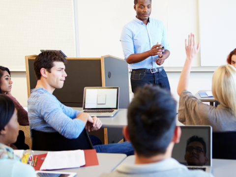 The image is of a Black professor standing in front of students, one student a women has her hand raised. 