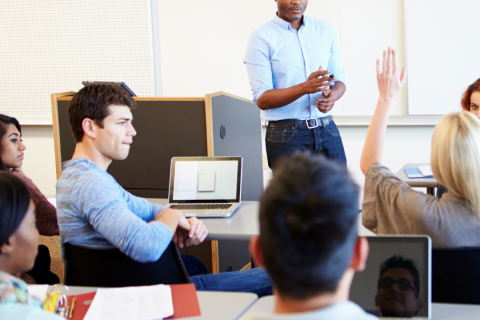 The image is of a Black professor standing in front of students, one student a women has her hand raised. 