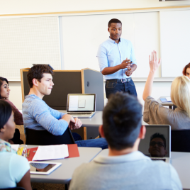 The image is of a Black professor standing in front of students, one student a women has her hand raised. 